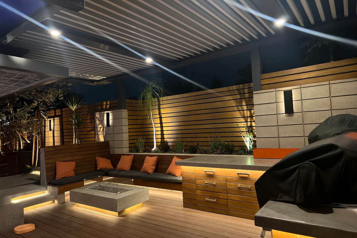 A modern outdoor patio with built-in seating, a fire pit, and wood paneling illuminated by warm accent lighting at night.